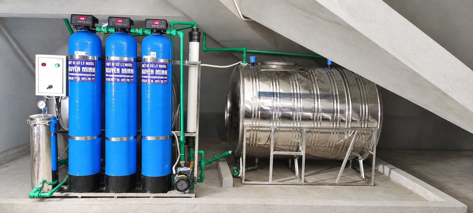 Hệ thống lọc nước với ba cột màu xanh và bồn inox bố trí dưới cầu thang